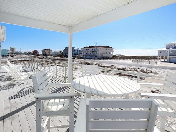 A covered outdoor seating area with white chairs and tables, overlooking a beach with buildings in the background.