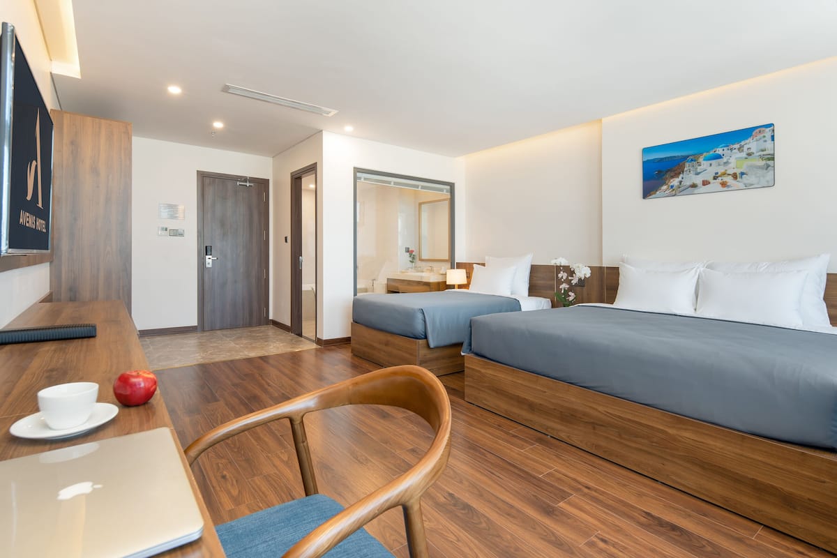 Habitación de hotel con piso de madera, un escritorio con una laptop y una manzana roja, dos camas con ropa de cama azul, un espejo grande y un cuadro de una escena costera.