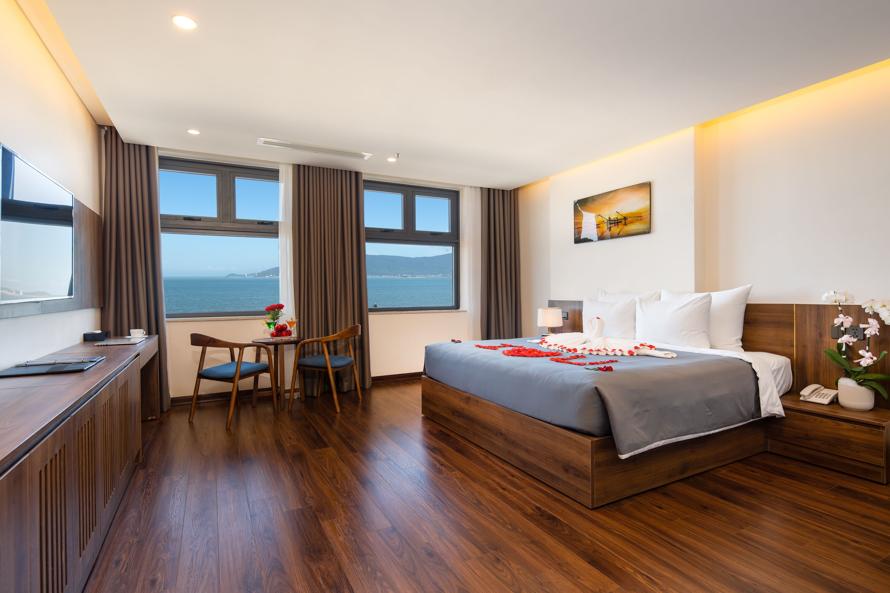 A hotel room with a large bed, a desk, and a chair. There is a window with a view of the sea.