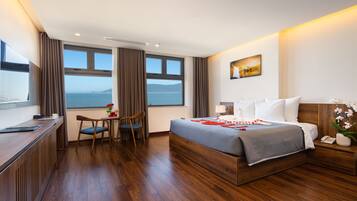 A hotel room with a large bed, a desk, and a chair. There is a window with a view of the sea.