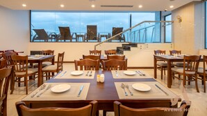 Un comedor con varias mesas y sillas, ventanas amplias con vista a sillas al aire libre, y una escalera al fondo.