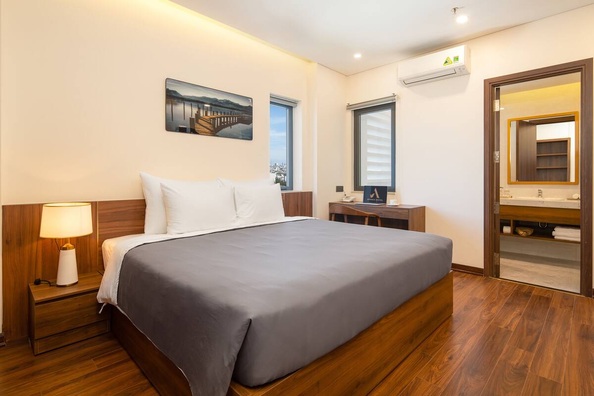 Una habitación de hotel moderna con una cama grande, una mesita de noche con lámpara, un baño con espejo y lavamanos, y un cuadro en la pared.