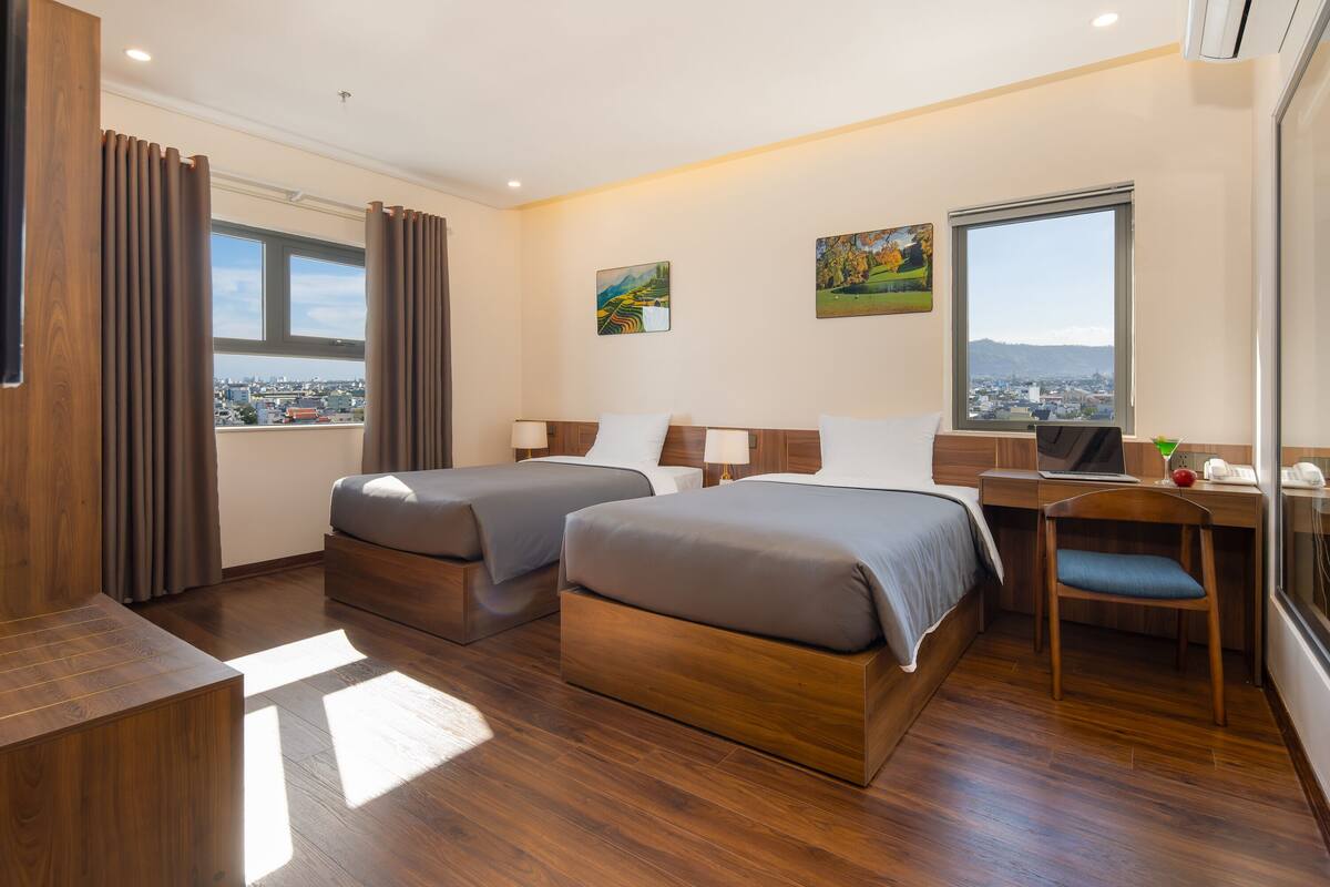 Habitación de hotel con dos camas, un escritorio, una silla y un ventanal grande con vistas a la ciudad.