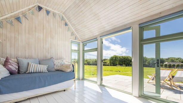 Une pièce chaleureuse avec un plafond en bois, un canapé bleu agrémenté de coussins à motifs et de grandes portes vitrées donnant sur une terrasse extérieure d’où l’on peut admirer un champ verdoyant et des éoliennes.