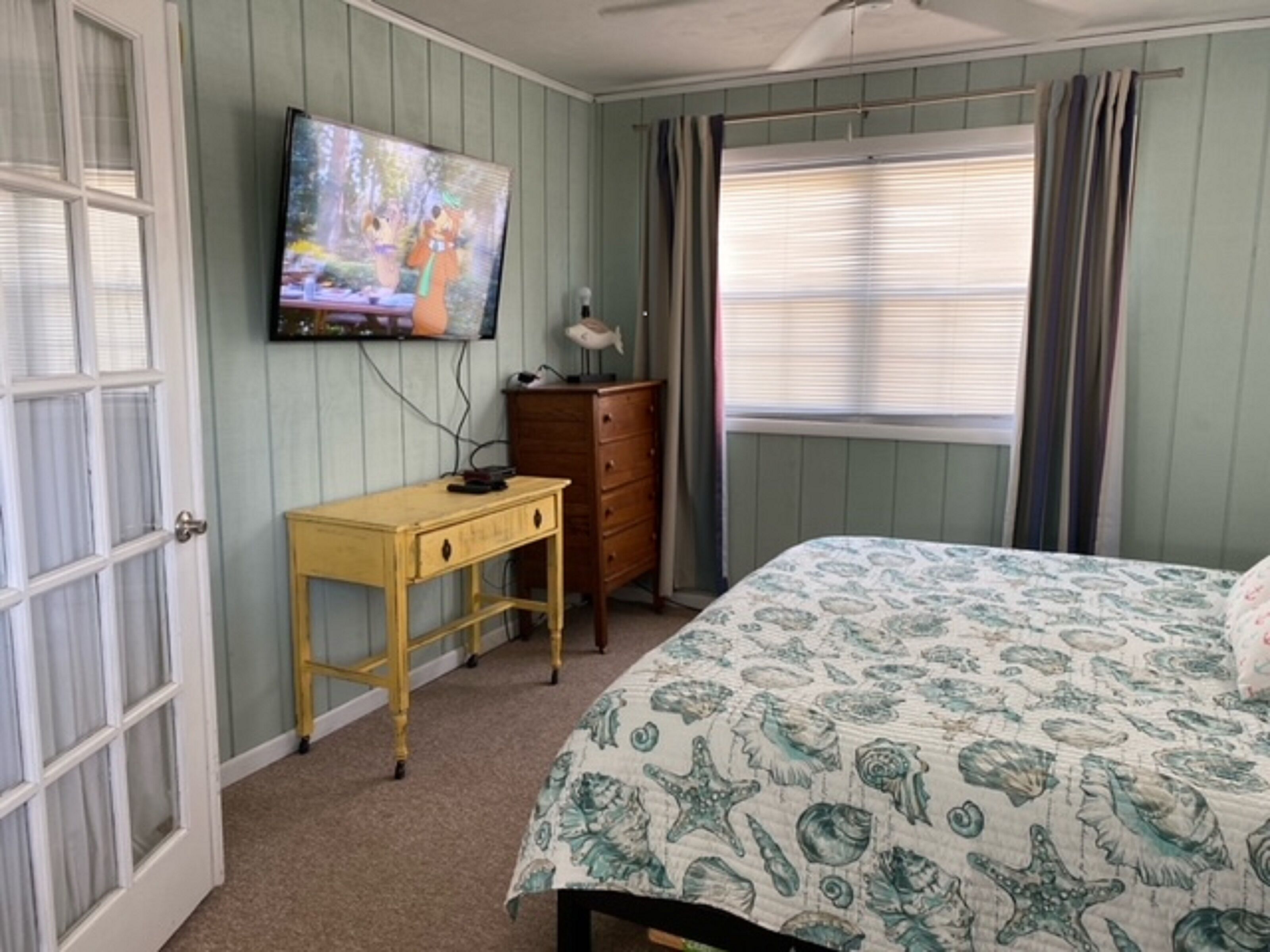 Quarto com cama, penteadeira, escrivaninha, televisão na parede e janela com cortinas.