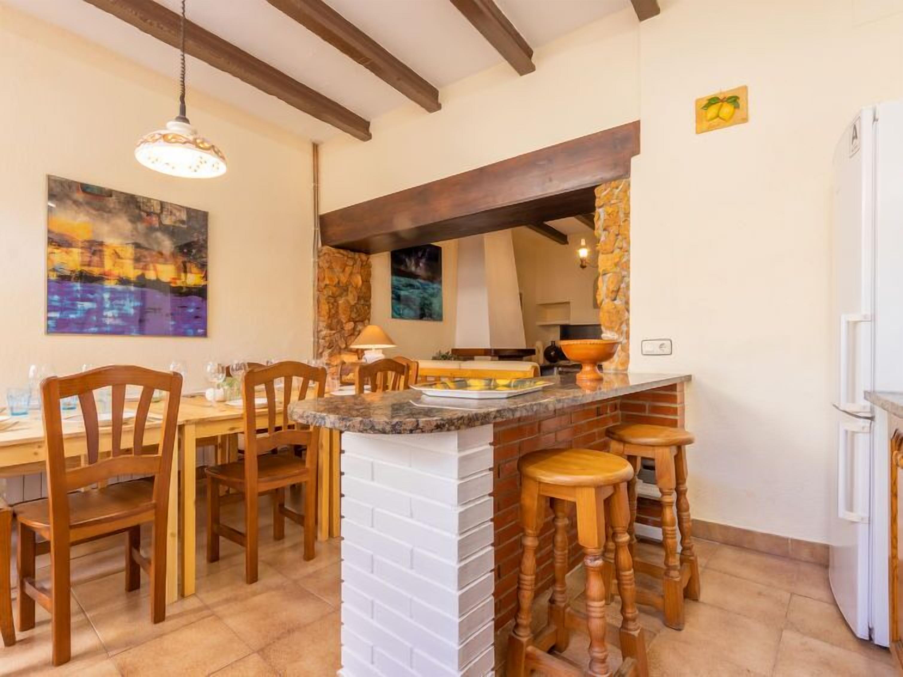 Un comedor con sillas de madera y una mesa, una cocina con pared de piedra, y una lámpara colgante.