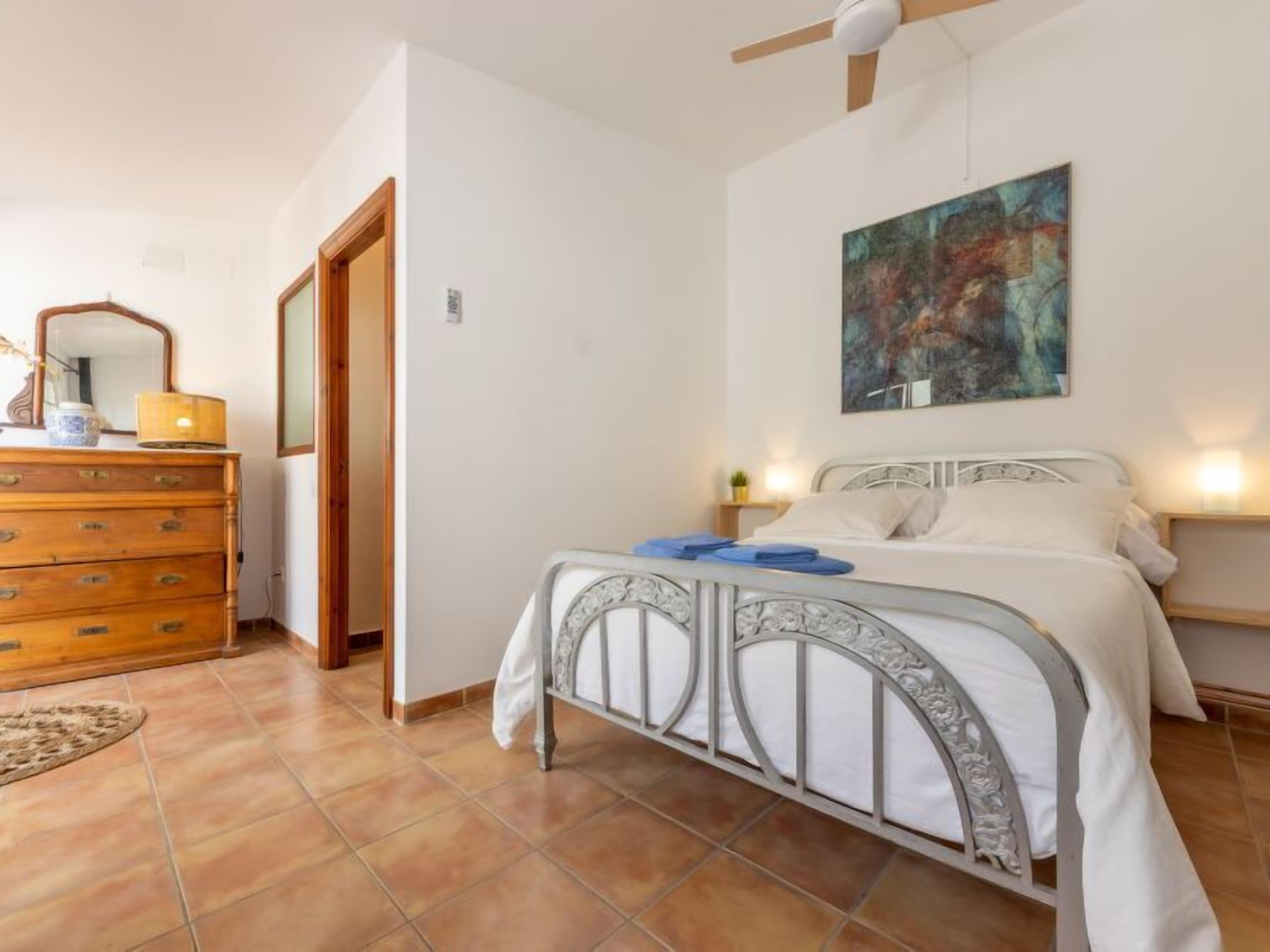 Una camera da letto con un letto a struttura in metallo, una cassettiera in legno, un quadro appeso al muro e una ventola a soffitto.