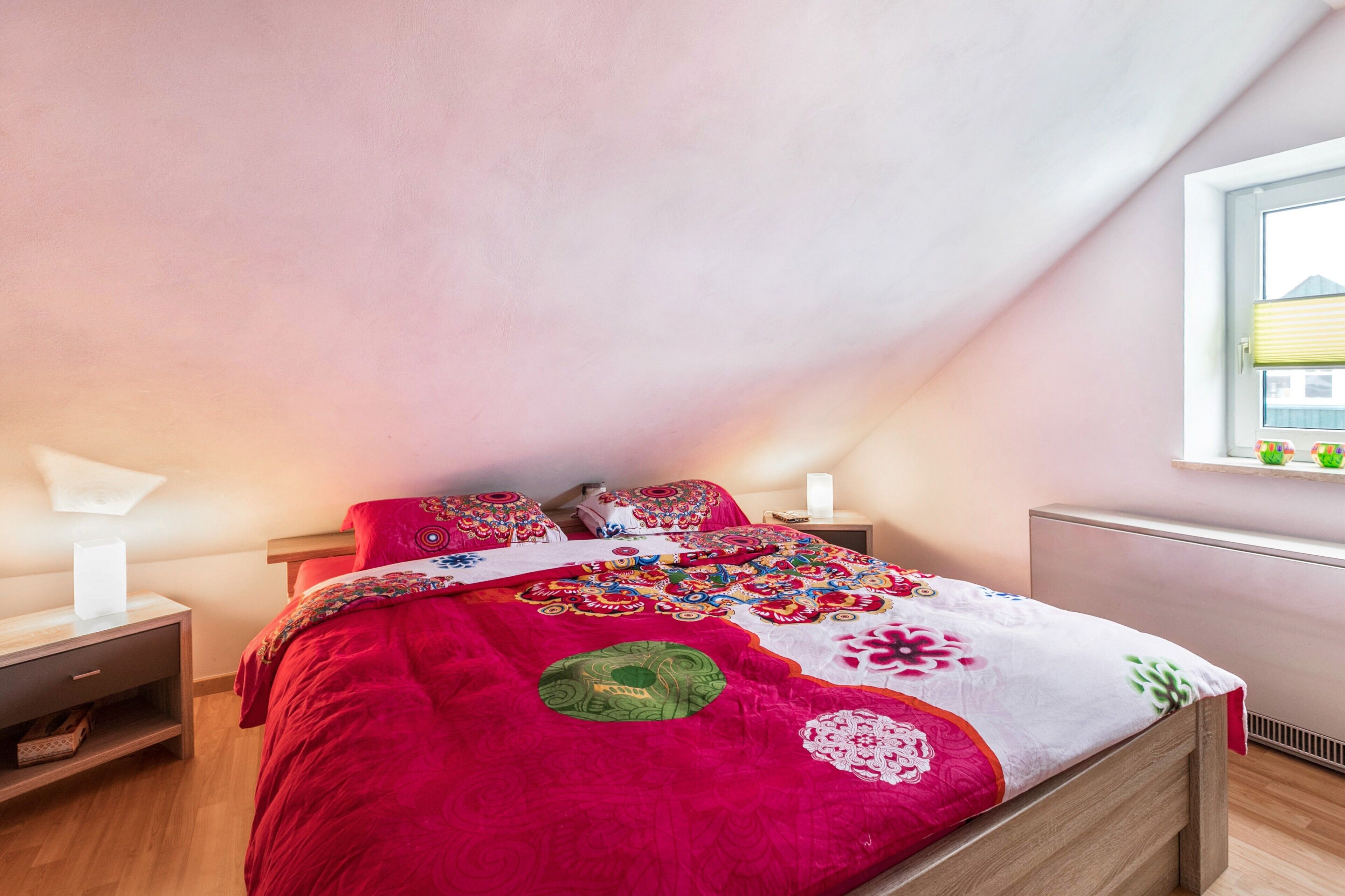 Un dormitorio con techo inclinado, una cama con colcha colorida, lámparas de noche y una ventana con persianas.