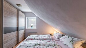 Un dormitorio con techo inclinado, una ventana con vistas, una cama con ropa de cama floral y una mesita de noche con lámpara.