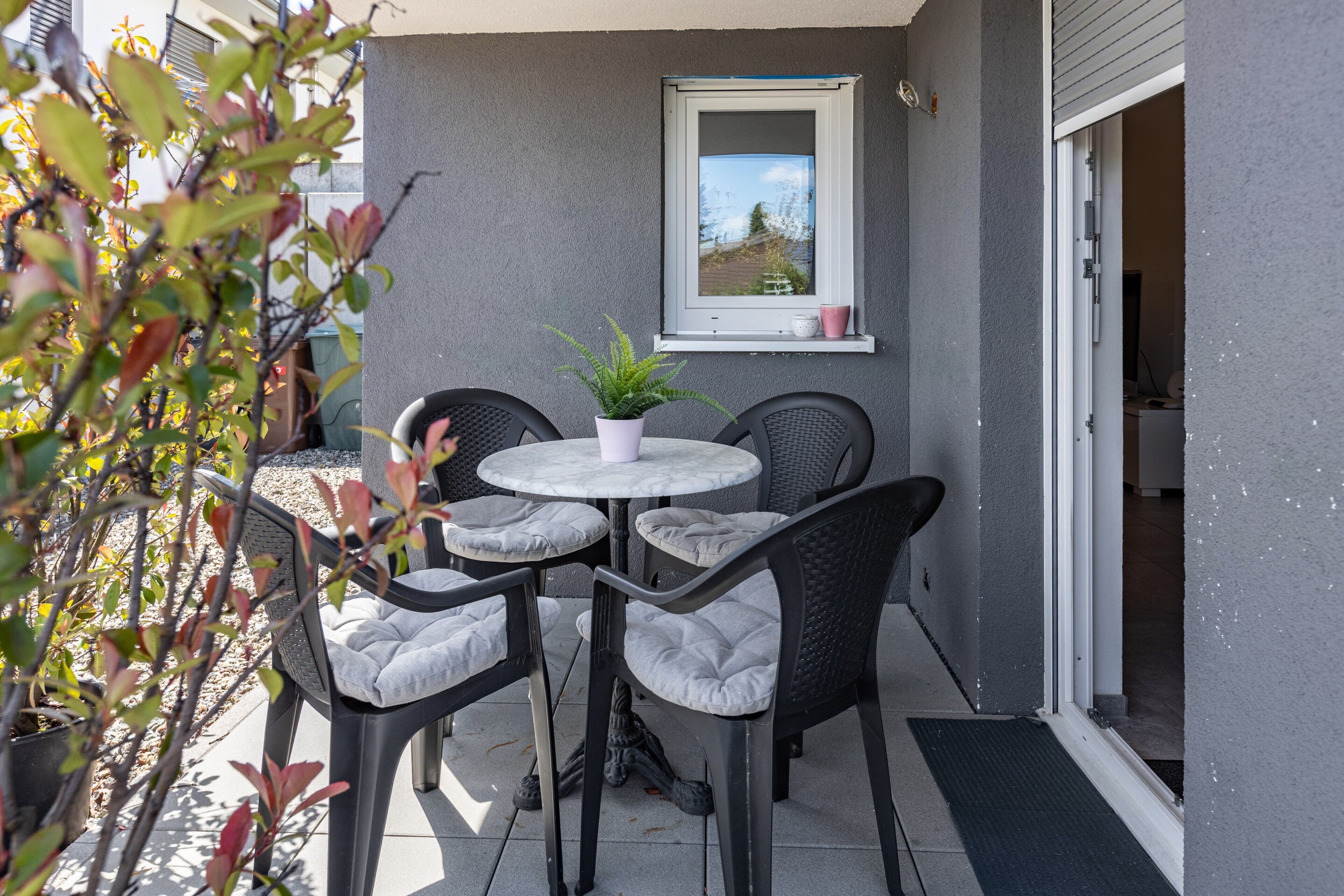An outdoor patio with four chairs, a table, a potted plant, and a window.