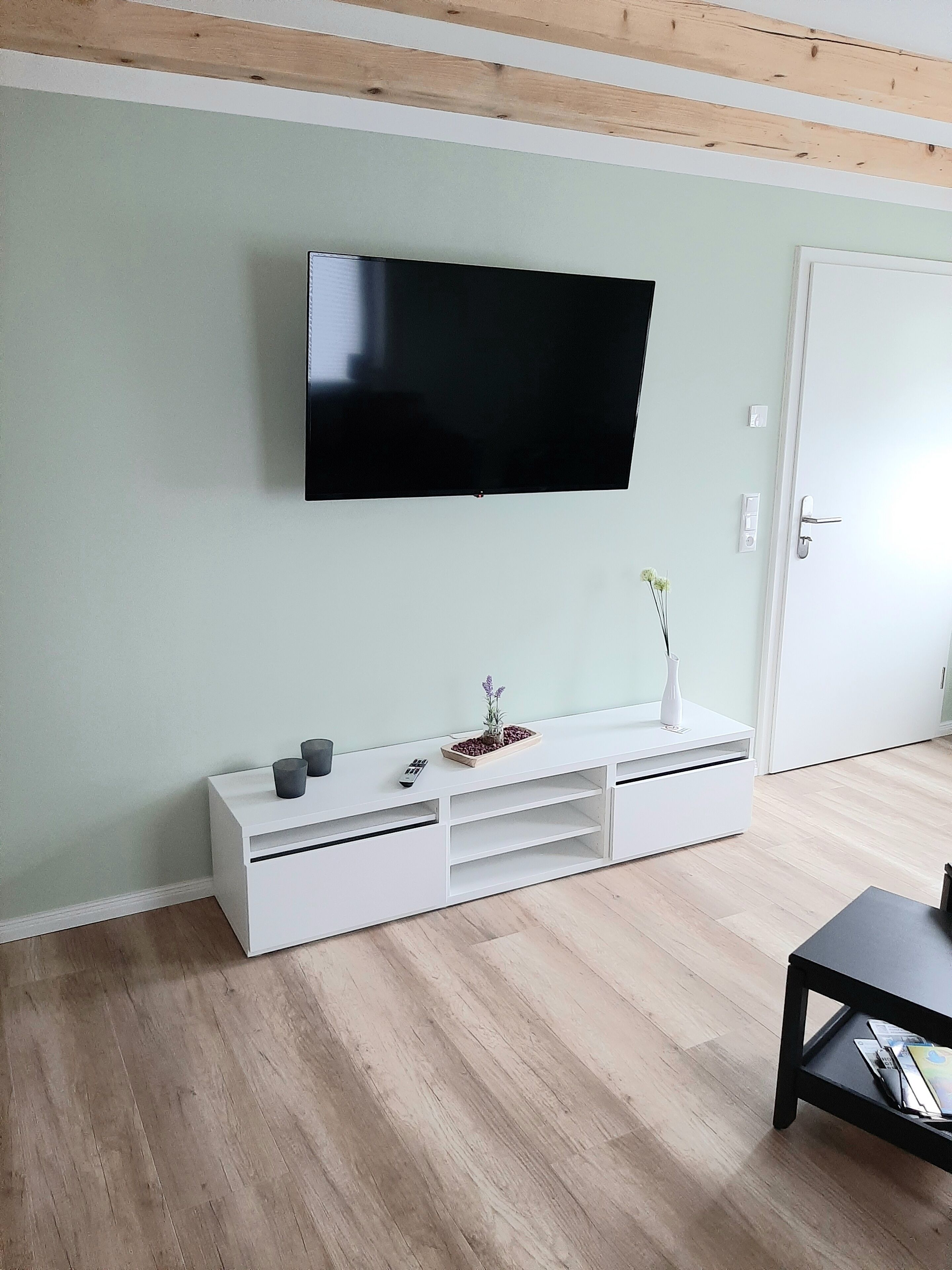 A modern living room with a flat-screen TV mounted on the wall, a white entertainment unit with a vase and remote controls, and a wooden floor.