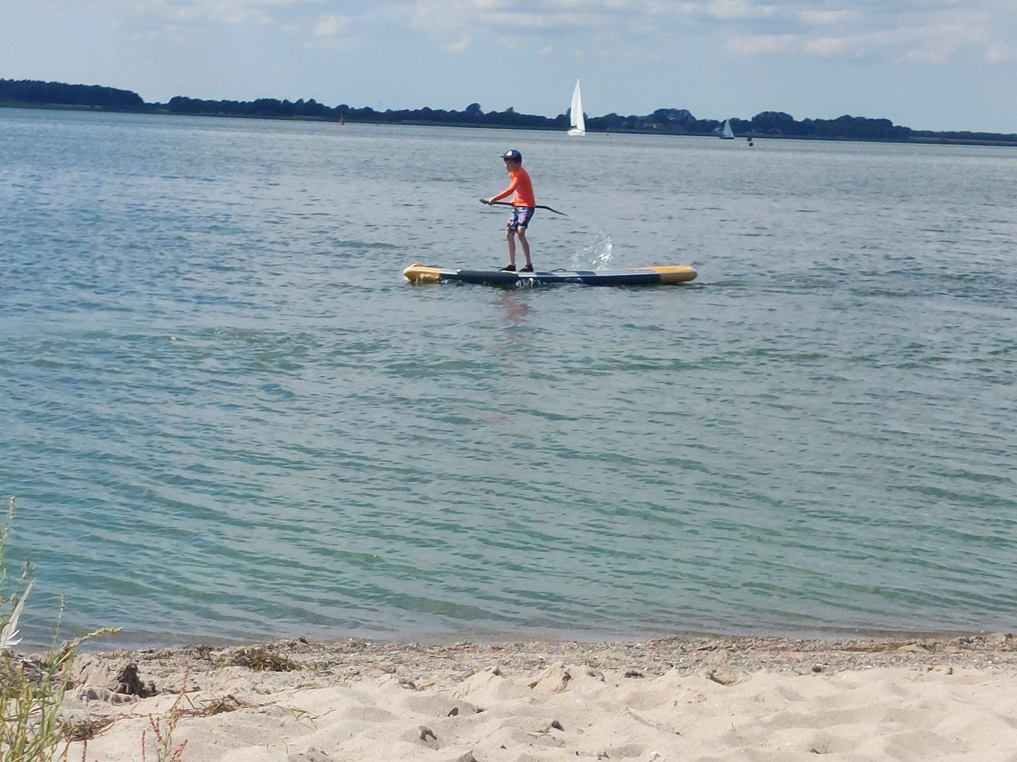 A person paddleboarding on a calm body of water with a sandy beach in the foreground and a distant sailboat on the horizon.