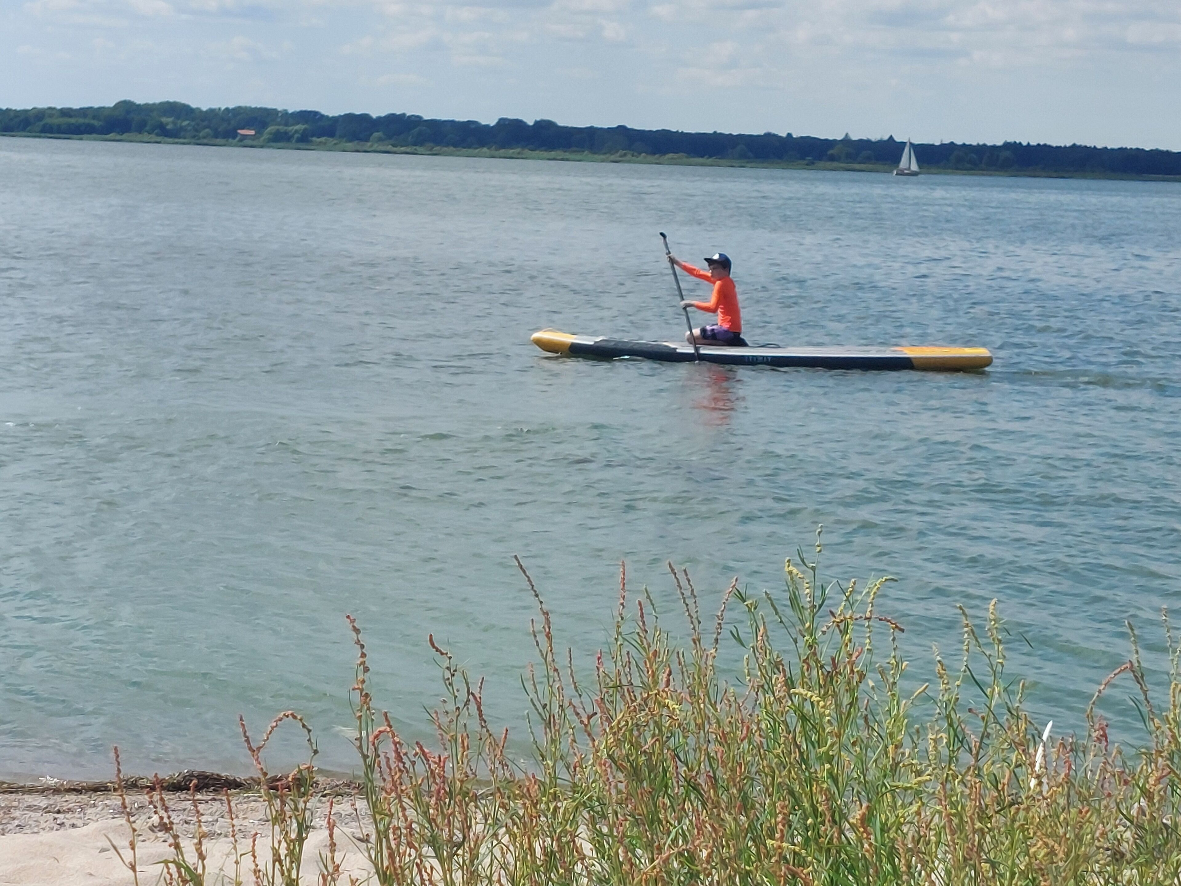 A person in a canoe on a calm body of water with a distant sailboat.