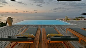 An infinity pool with a wooden deck, lounge chairs, and a large vase. - Villa Luna 8 by Massimo Villas (Cefalù)