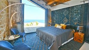 Een slaapkamer met een groot bed, uitzicht op de oceaan, een klein tafeltje met een kopje en boek, en een stoel.