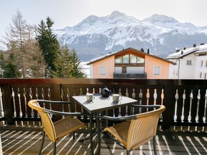 Un balcon avec des fauteuils en osier et une table, d’où l’on peut admirer des montagnes enneigées et une maison en bois.