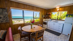 Ein gemütlicher Raum mit Holzvertäfelung, einem runden Esstisch mit Stühlen, einer Küchenzeile mit Kühlschrank und großen Fenstern mit Blick auf den Strand und Palmen.
