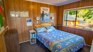 Ein gemütliches Schlafzimmer mit einer Holzwand, einem Bett mit floralem Überwurf, einem Bücherregal und einem Fenster mit Blick auf Grünflächen.