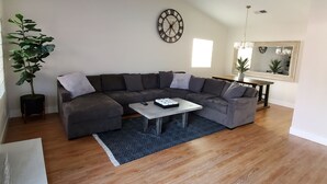 Uma sala de estar moderna com um amplo sofá em formato de L, uma mesa de centro, um vaso de planta e um relógio de parede.