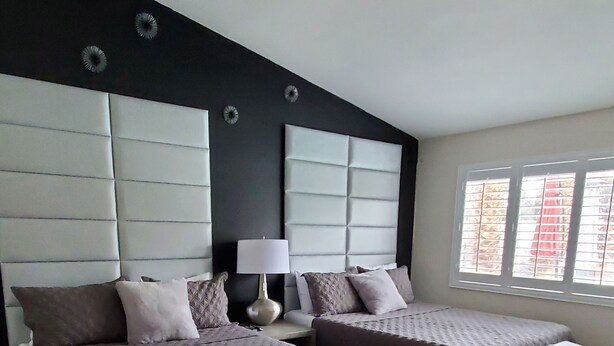 Quarto moderno com duas camas, uma cabeceira grande e acolchoada, mesa de cabeceira com luminária e janela com persianas.