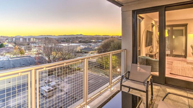 A balcony with a view of a cityscape at sunset, a glass door leading to a room with a sofa and a lamp. - Luxury Condo, Downtown Asheville (Asheville)
