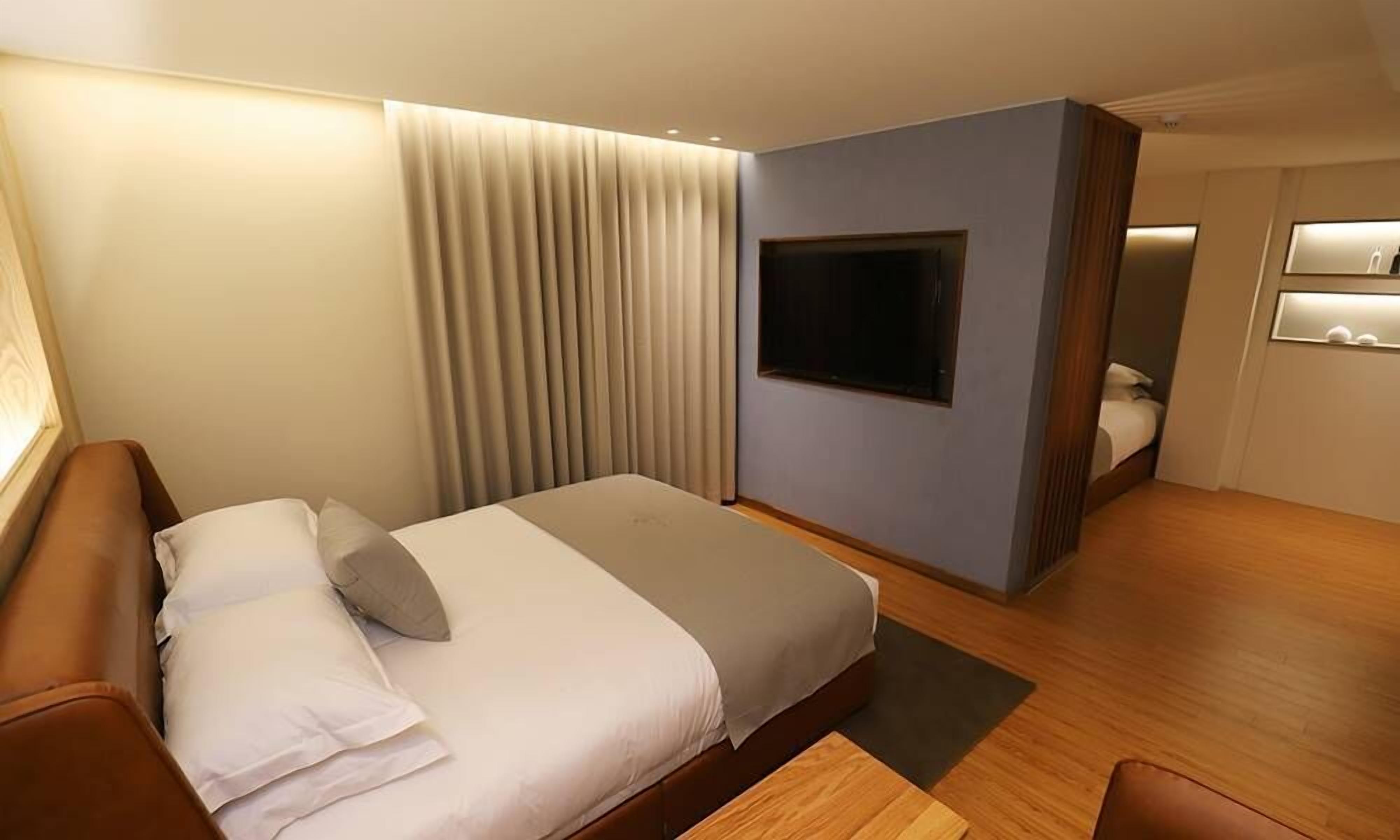 A modern hotel room with a bed, a TV, a mirror, and a small seating area.