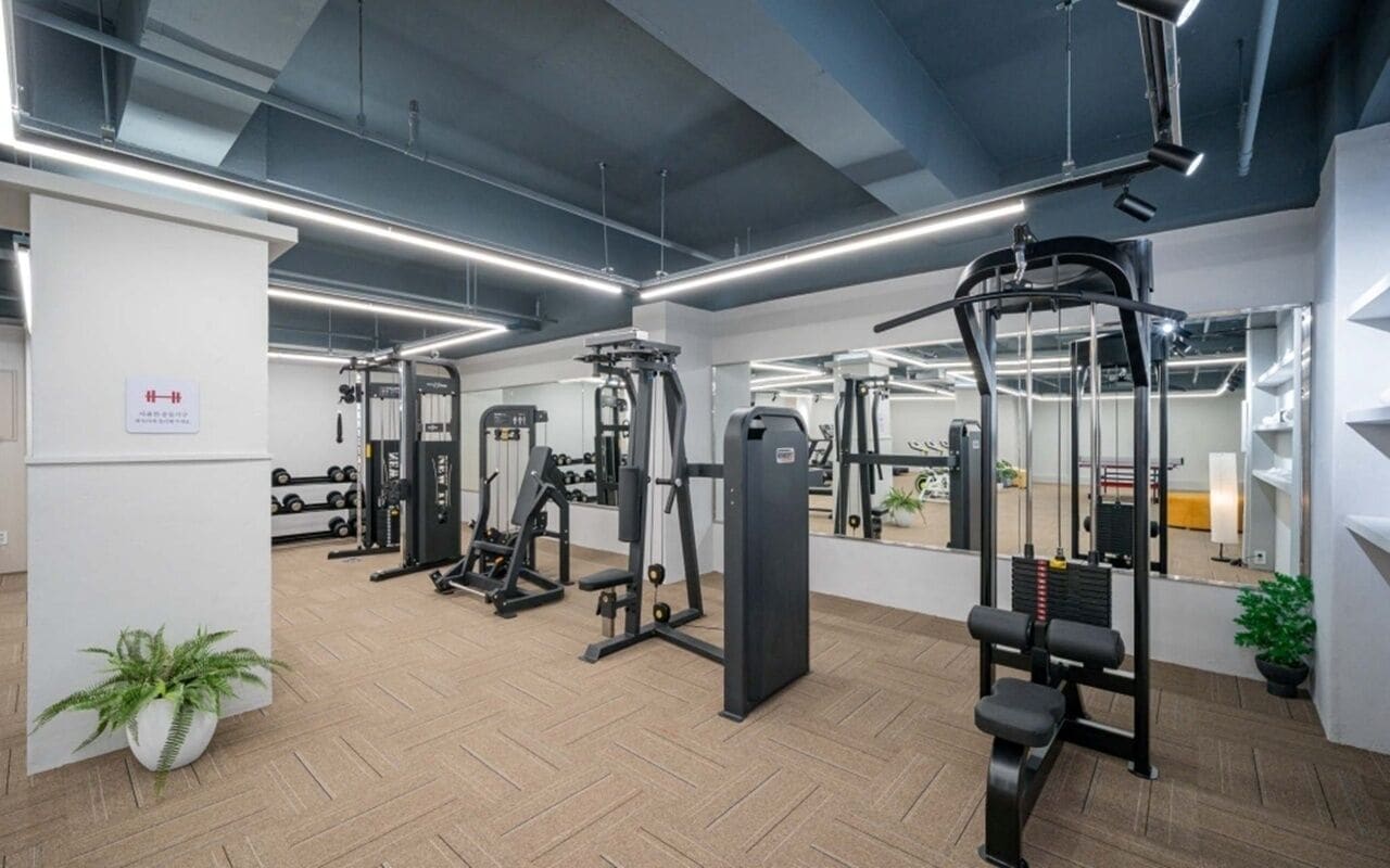 Uma academia com diversos aparelhos de exercício, um pilar branco com uma placa e um vaso de planta.