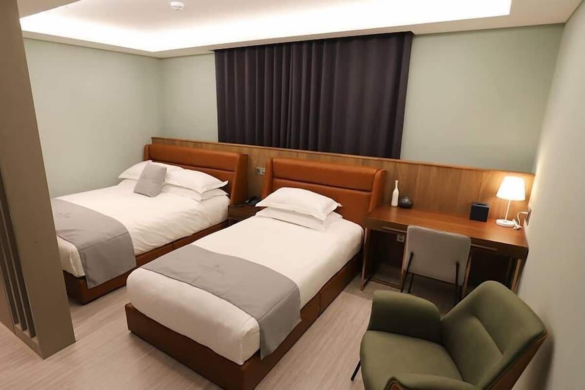 A hotel room with two beds, a desk, a chair, and a lamp.