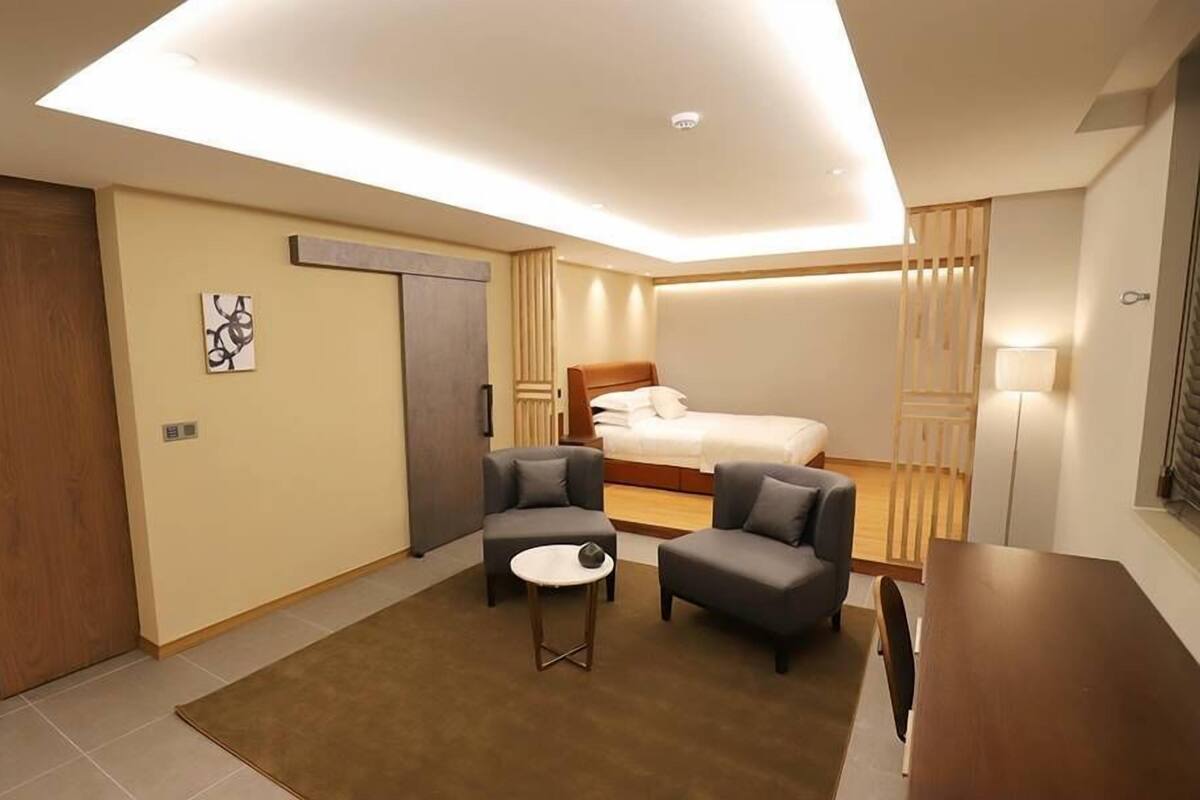 A modern hotel room with a bed, two armchairs, a small table, and a ceiling light.