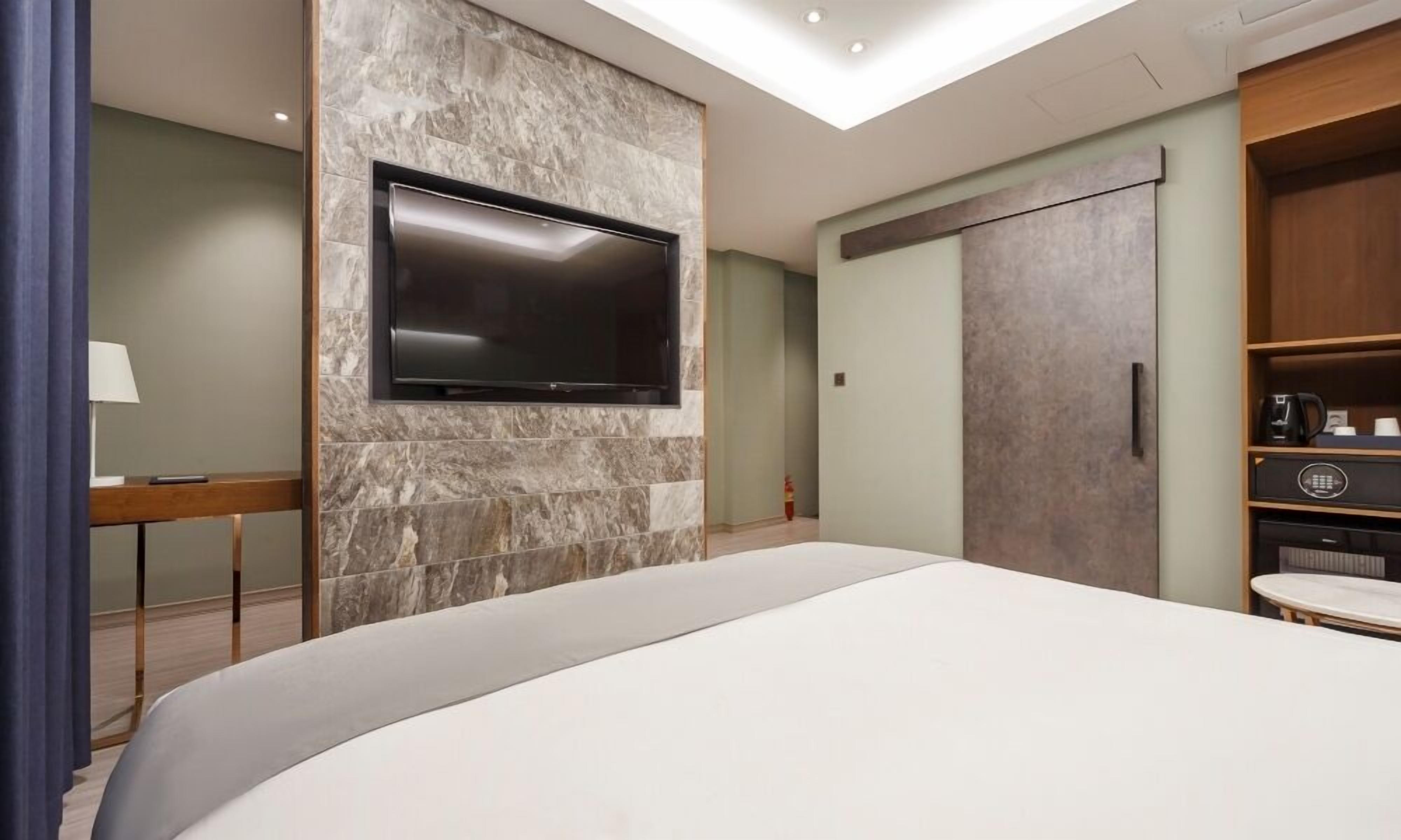 A modern hotel room with a large bed, a flat-screen TV mounted on a stone wall, a wardrobe, and a small table with a lamp.