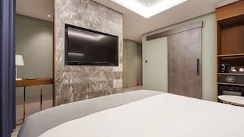 A modern hotel room with a large bed, a flat-screen TV mounted on a stone wall, a wardrobe, and a small table with a lamp.