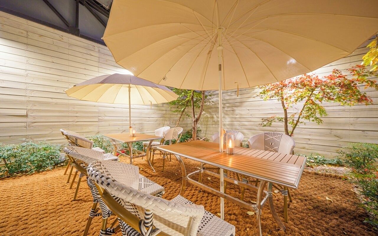Un patio al aire libre con una mesa y sillas de madera, un paraguas grande y un árbol pequeño.