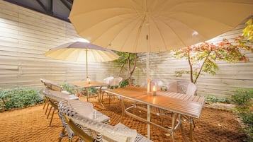 Un patio al aire libre con una mesa y sillas de madera, un paraguas grande y un árbol pequeño.