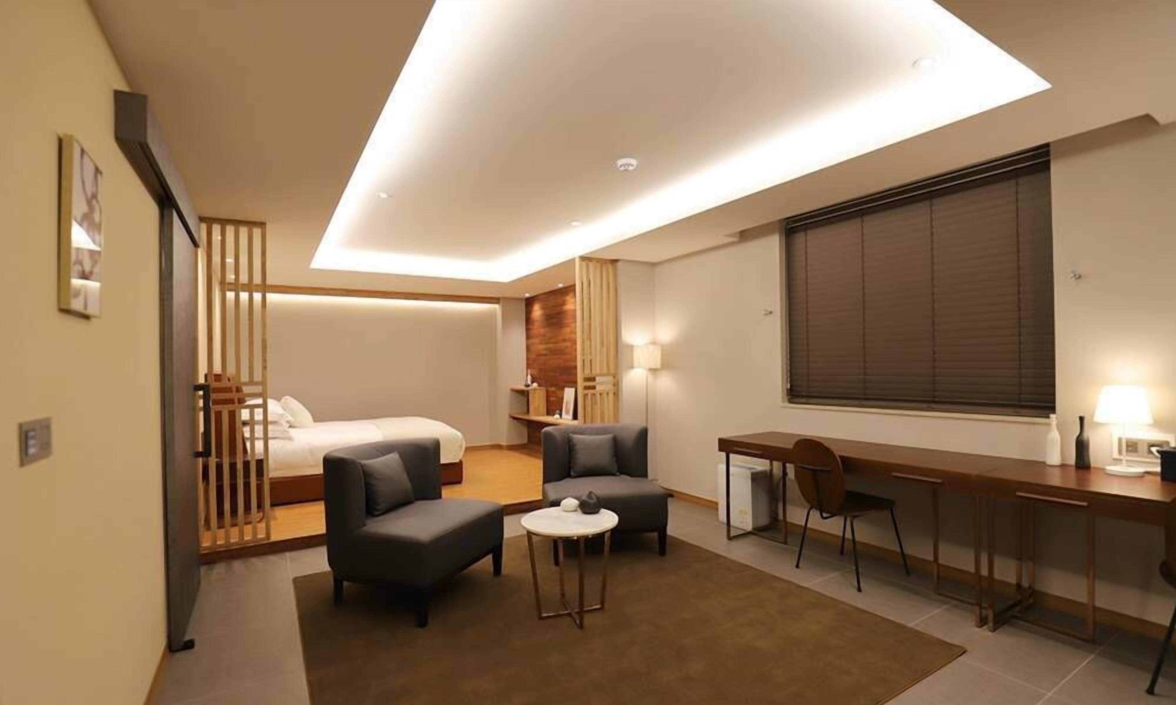 A modern hotel room with a bed, two armchairs, a small round table, a desk with a chair, a large window with blinds, and a ceiling light.