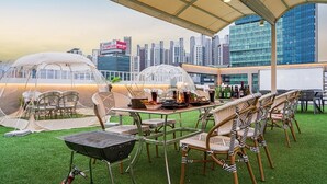 An outdoor dining area with transparent canopies, wicker chairs, a grill, and a city skyline view. - Dongdaegu Station Eastern Hotel (Daegu)