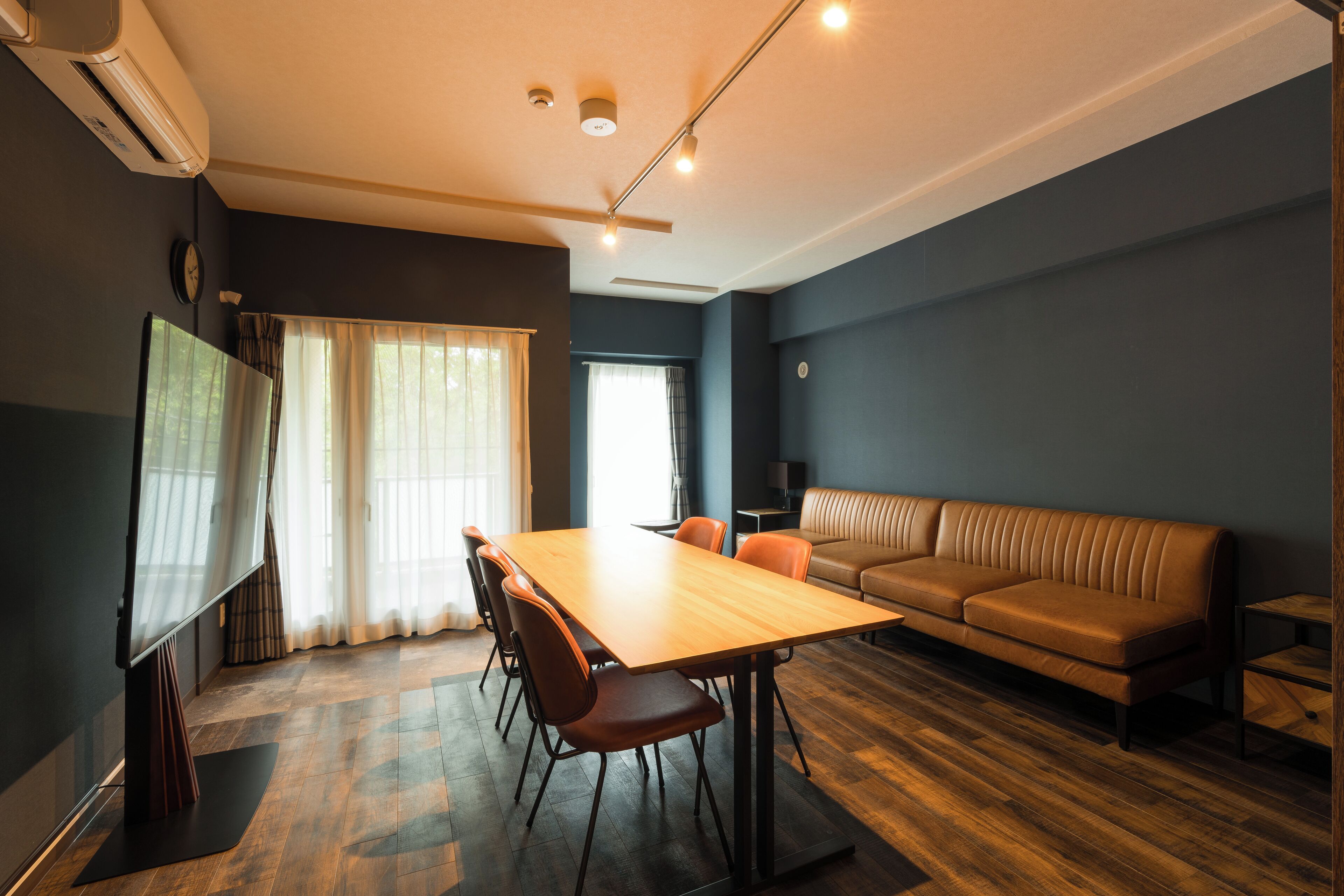 A modern living room with a wooden dining table, orange chairs, a brown leather sofa, and a flat-screen TV.