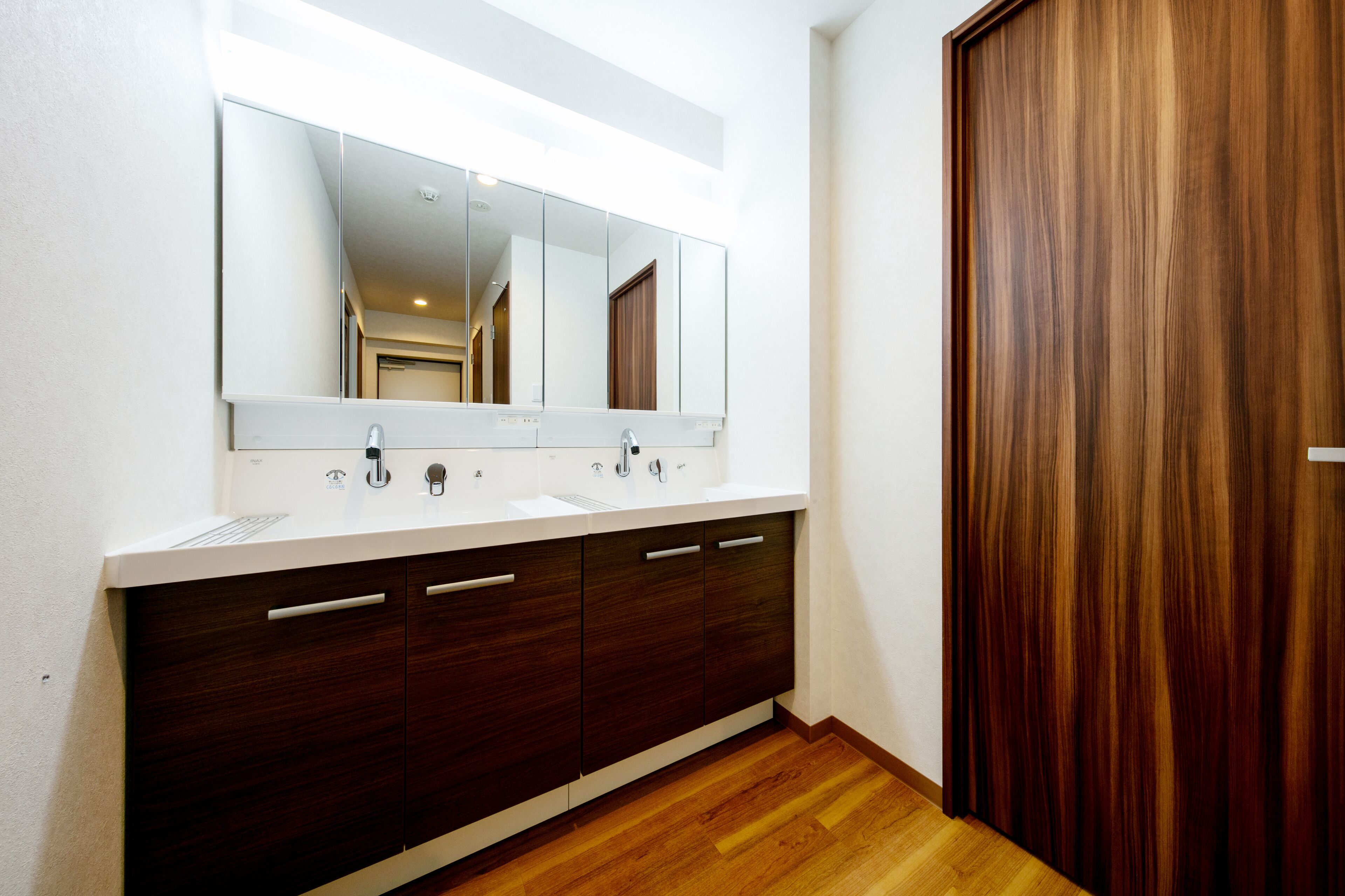 Un baño moderno con un espejo grande, dos lavamanos, armarios de madera oscura y una puerta de madera.