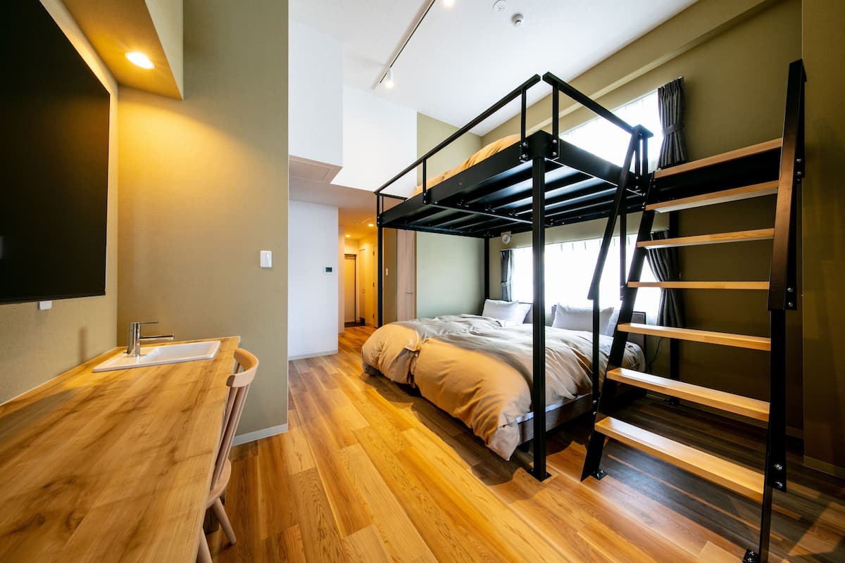 A bedroom with a bunk bed, wooden flooring, a small bathroom with a sink, and a window with curtains.
