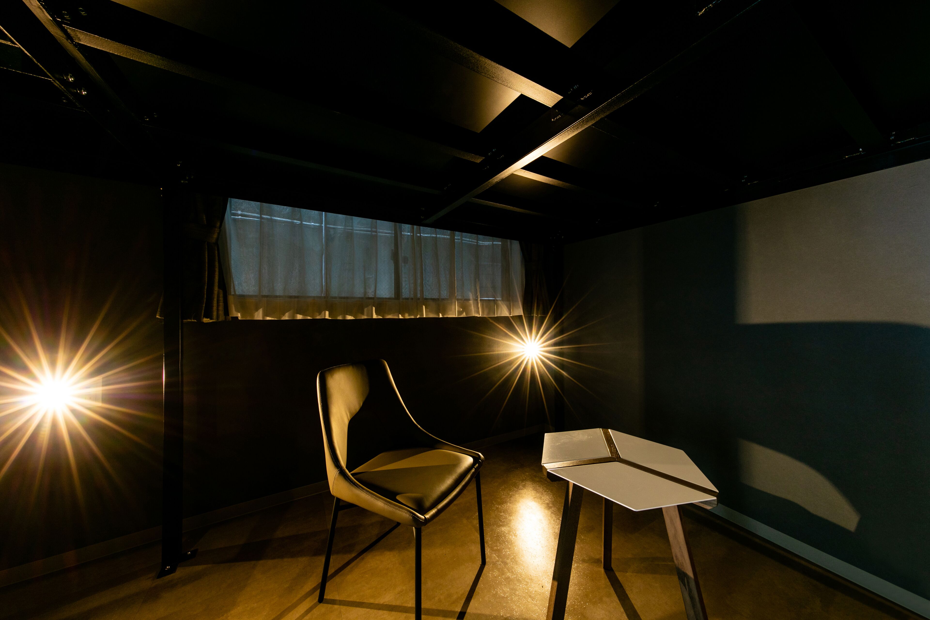 Una habitación con poca luz, una silla y una pequeña mesa, una ventana con cortinas translúcidas, y un techo con vigas y luminarias a la vista.