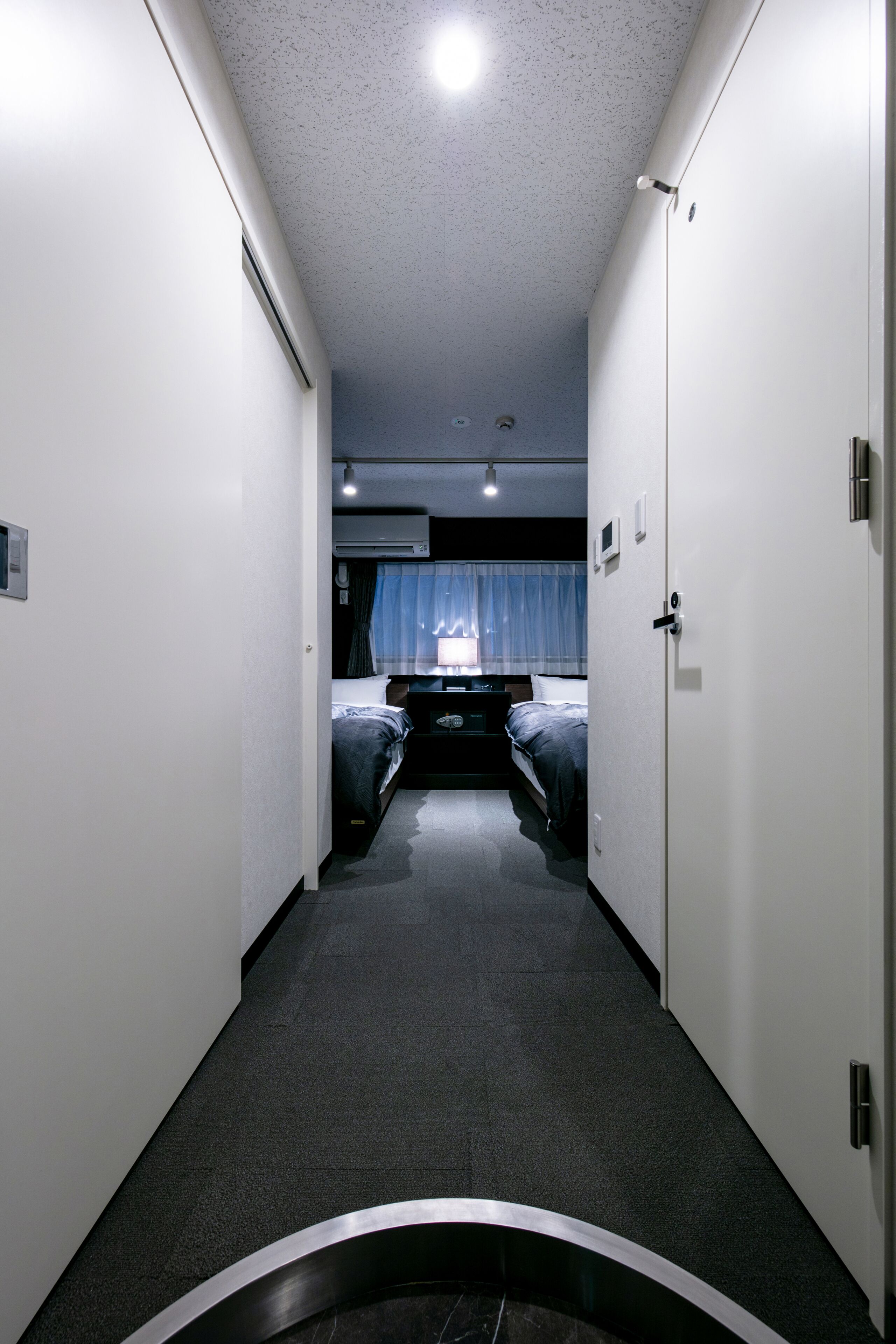 Un couloir étroit mène à une chambre à coucher qui contient un lit, une table de chevet et une fenêtre avec des rideaux.