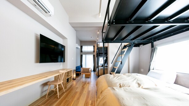 Una habitación de hotel moderna con suelo de madera, una cama, un escritorio con silla, un televisor montado en la pared y una escalera que lleva a un nivel superior.