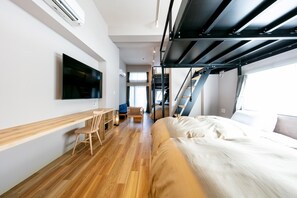 A modern hotel room with a wooden floor, a bed, a desk with a chair, a TV mounted on the wall, and a staircase leading to an upper level.