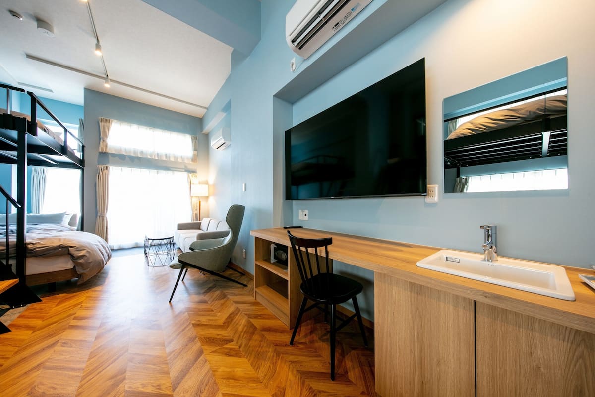 A modern hotel room with a wooden herringbone floor, a bed with a grey headboard, a desk with a sink, a large flat-screen TV, and a window with curtains.
