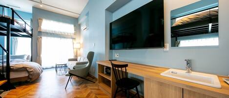 Una habitación de hotel moderna con suelo de madera en espiga, una cama con cabecera gris, un escritorio con lavabo, un televisor de pantalla plana grande y una ventana con cortinas.