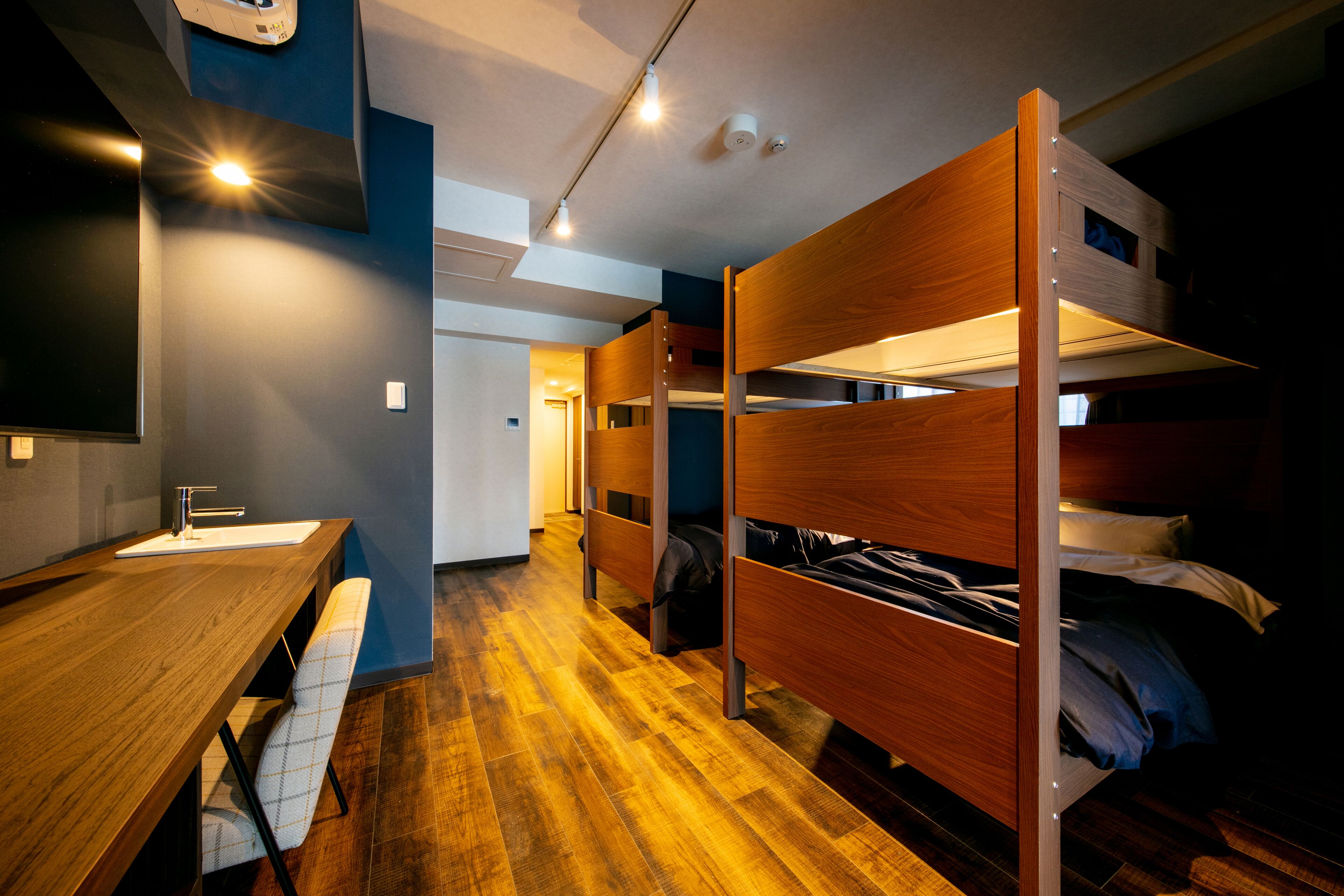Een slaapkamer met een stapelbed, een houten bureau en bankje, een wastafel en een gang die naar een andere kamer leidt.