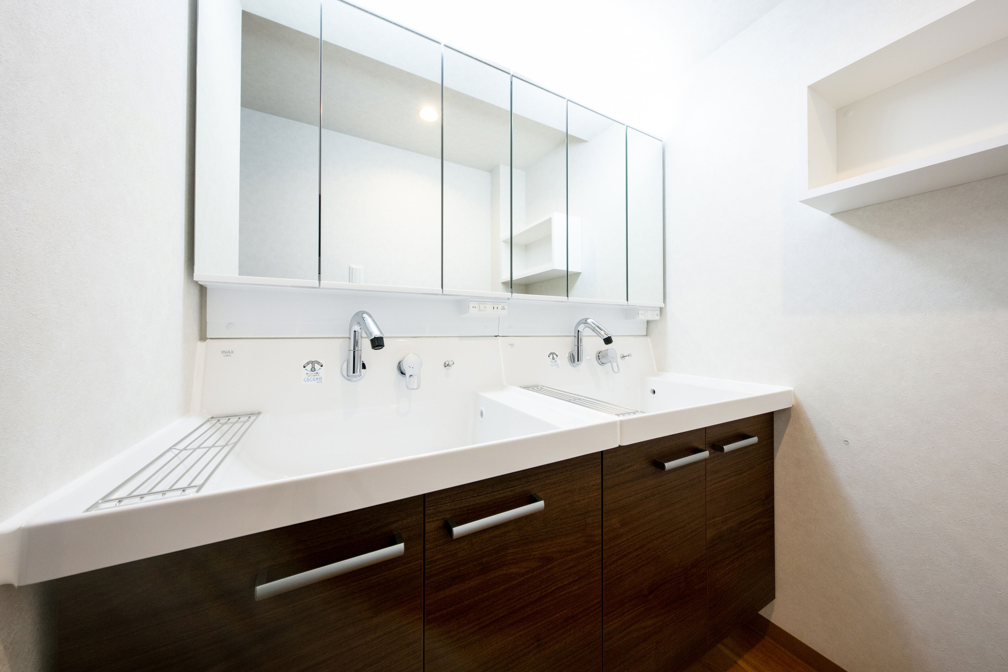A modern bathroom with a white sink, two faucets, and a large mirror above. There are wooden cabinets with silver handles and a white countertop.