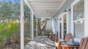 Un portico riparato con sedie e tavolo in legno, una ventola a soffitto e vista su alberi e un edificio sullo sfondo.