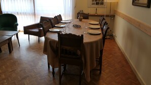A dining area with a round table set for six, surrounded by wooden chairs. A green armchair is visible to the left, and a radiator is on the wall to the right.