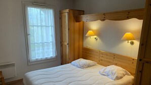 Ein Schlafzimmer mit einem hölzernen Kopfteil, zwei Nachttischlampen, einem Bett mit weißer Bettwäsche, einem Fenster mit durchsichtigen Vorhängen und einer Heizung.