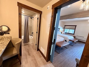 A hotel room with a bed, a sitting area with a bench, a small table with a chair, a shower area, and a view of the outside. - Arrowhead Properties Unit 7 (West Yellowstone)